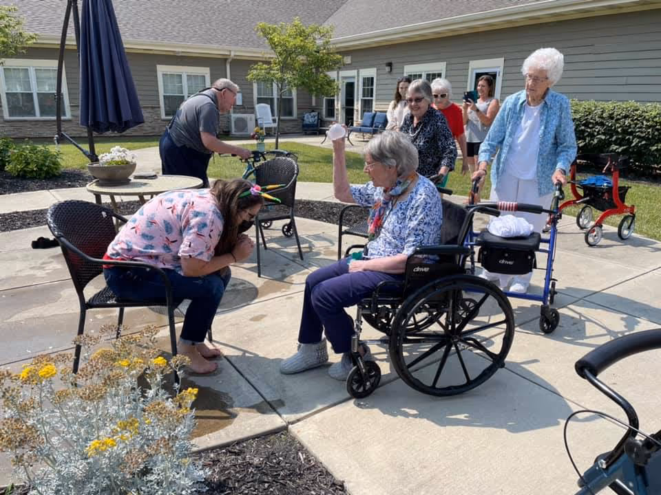A group of elderly people and a younger woman are gathered outside on a sunny day at a senior living facility. One elderly woman in a wheelchair is raising her hand while interacting with the younger woman who is sitting on a chair and leaning forward. Other elderly individuals are standing or using walkers nearby. The setting includes a paved patio area with chairs, plants, and a building with windows in the background.