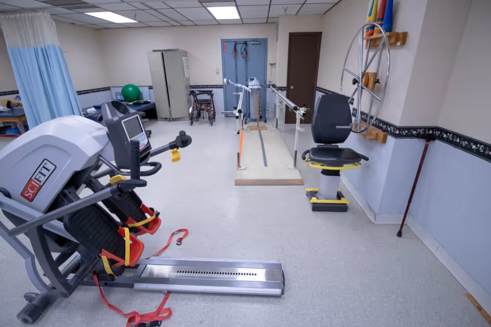 A physical therapy room with exercise equipment including a SciFit machine, parallel bars for walking practice, a seated exercise bike, a walker, and various therapy tools. The room has light-colored walls with a floral border and a tiled ceiling.