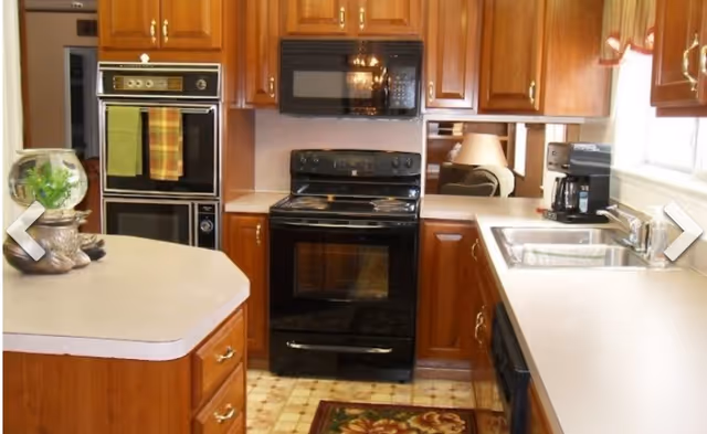 A kitchen with wooden cabinets, a black electric stove with oven, a black microwave above the stove, a double sink under a window with a coffee maker on the counter, and a small island with a decorative fishbowl and a ceramic fish figurine.