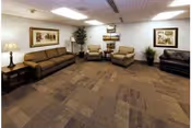 A spacious living room area with beige carpet flooring, a brown sofa, two beige armchairs, a black leather sofa, side tables with lamps, framed artwork on the walls, and potted plants in the corners.