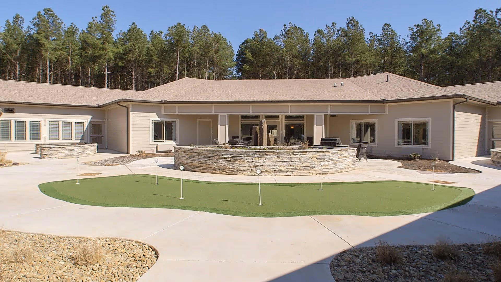 Outdoor courtyard area of a senior living facility with a putting green, circular stone fire pit, patio chairs, and a single-story building with beige siding and multiple windows surrounded by tall pine trees under a clear blue sky.