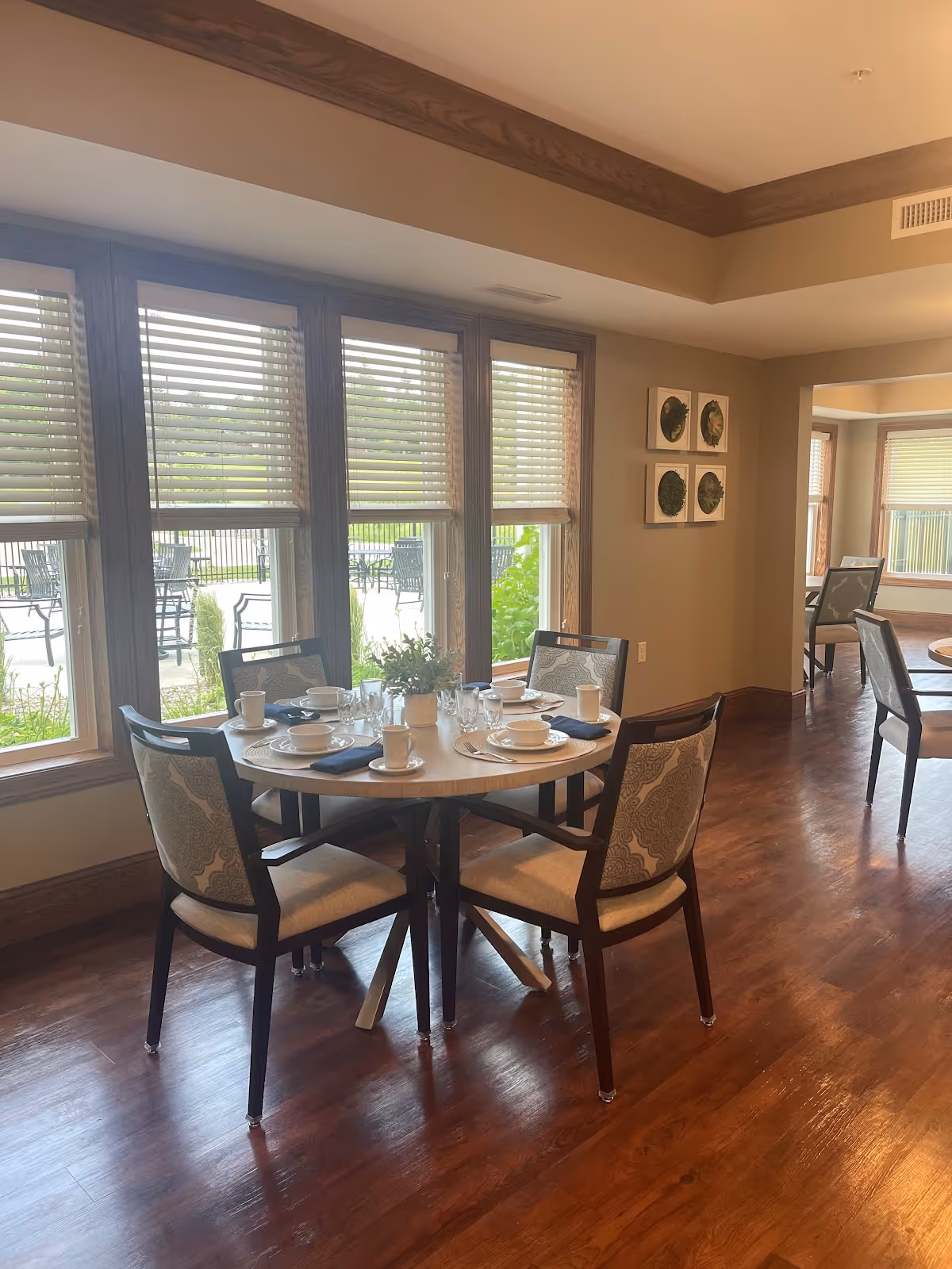 A dining area in a senior living facility with a round table set for four people. The table has white plates, cups, glasses, and dark napkins. Four cushioned chairs with patterned backs surround the table. Large windows with wooden frames and blinds let in natural light, showing an outdoor patio with tables and chairs. The room has wooden flooring and beige walls with decorative wall art.