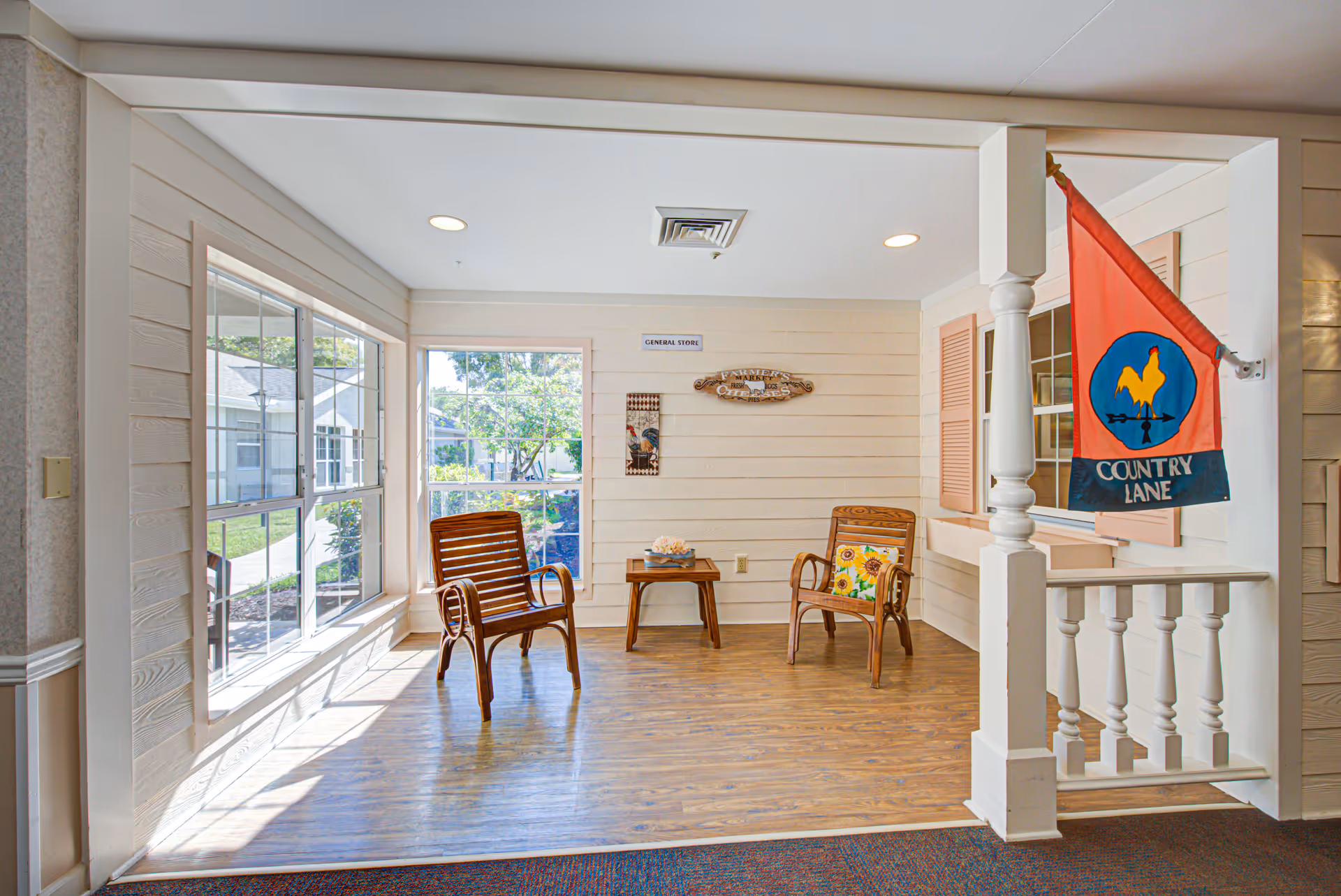 A bright indoor sitting area with two wooden chairs and a small wooden table between them. The walls are light-colored with horizontal paneling, and there are large windows letting in natural light. A decorative flag with a rooster and the words 'Country Lane' hangs on a white post. Wall signs include one that says 'GENERAL STORE' and another with a rooster design.