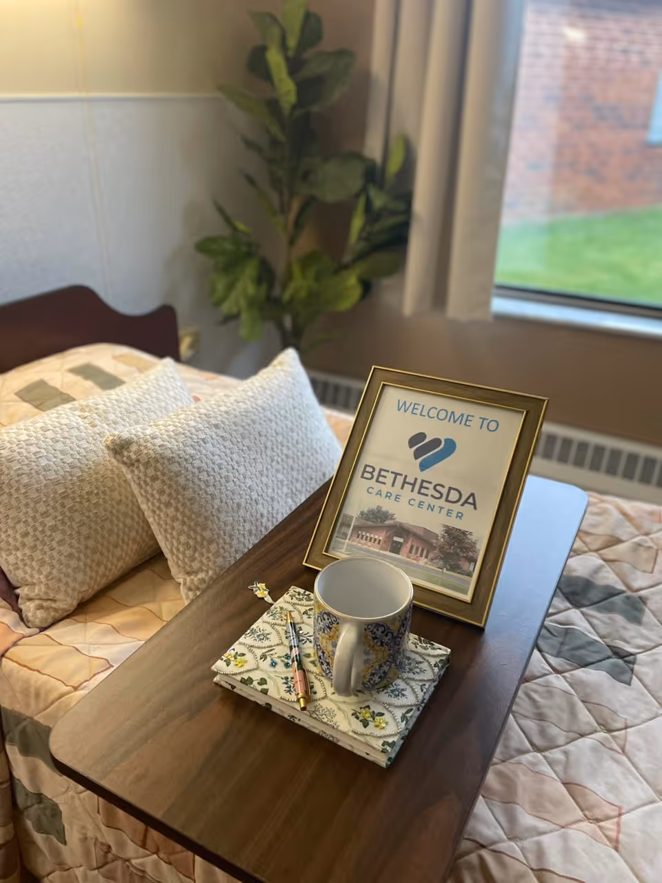 A cozy bedroom setup in Bethesda Care Center featuring a bed with two textured pillows and a quilted blanket. On a wooden overbed table, there is a framed welcome sign for Bethesda Care Center, a decorative mug, a floral napkin, and a pen. A green leafy plant is visible in the background near a window with beige curtains.
