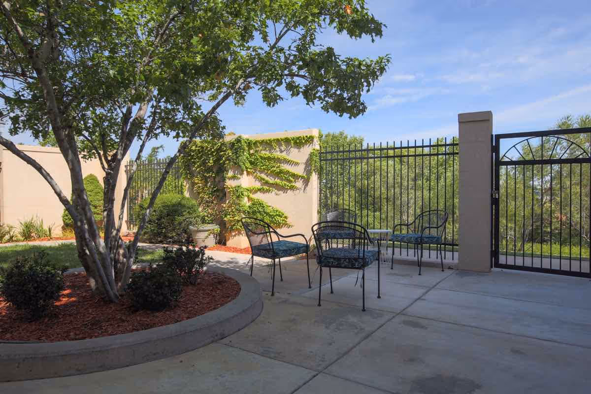 Outdoor patio area with metal chairs and a small table on a concrete surface, surrounded by a low wall and black metal fence. There is a tree and some bushes planted in a circular raised bed with red mulch, and a clear blue sky overhead.
