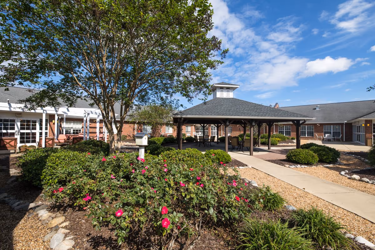 Outdoor courtyard area at Elizabeth City Health & Rehabilitation featuring a large tree, flowering bushes, a covered pavilion with seating, and a paved walkway surrounded by landscaping and garden beds under a partly cloudy blue sky.