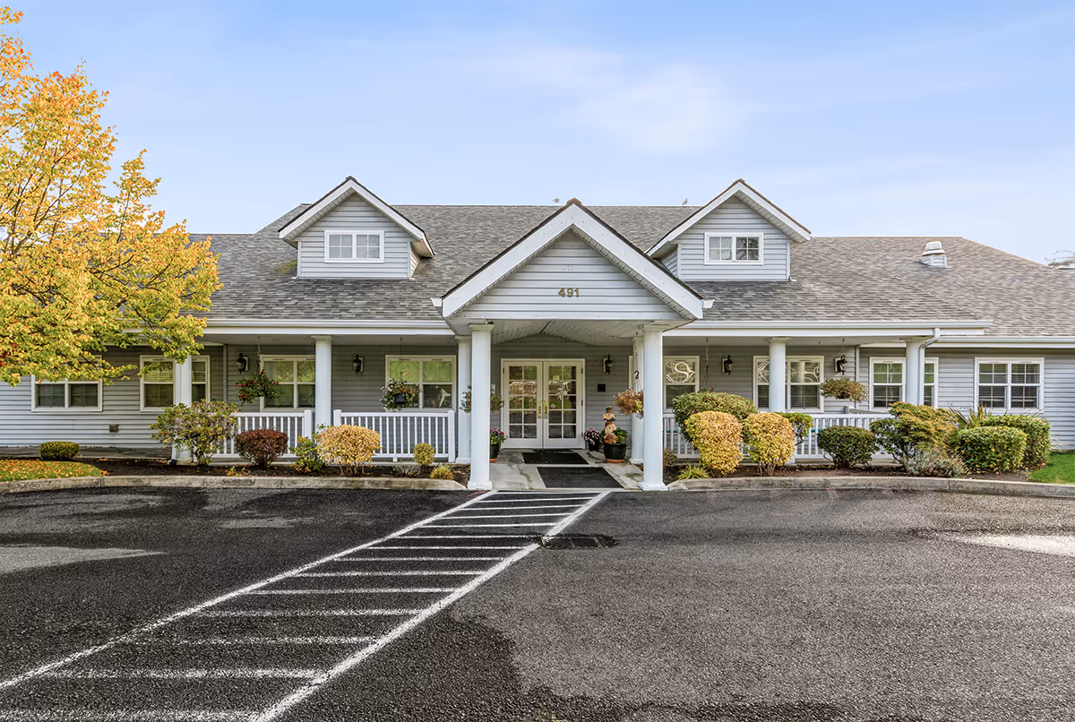 Front entrance of a single-story senior living building with a covered porch, columns, and landscaped shrubs.