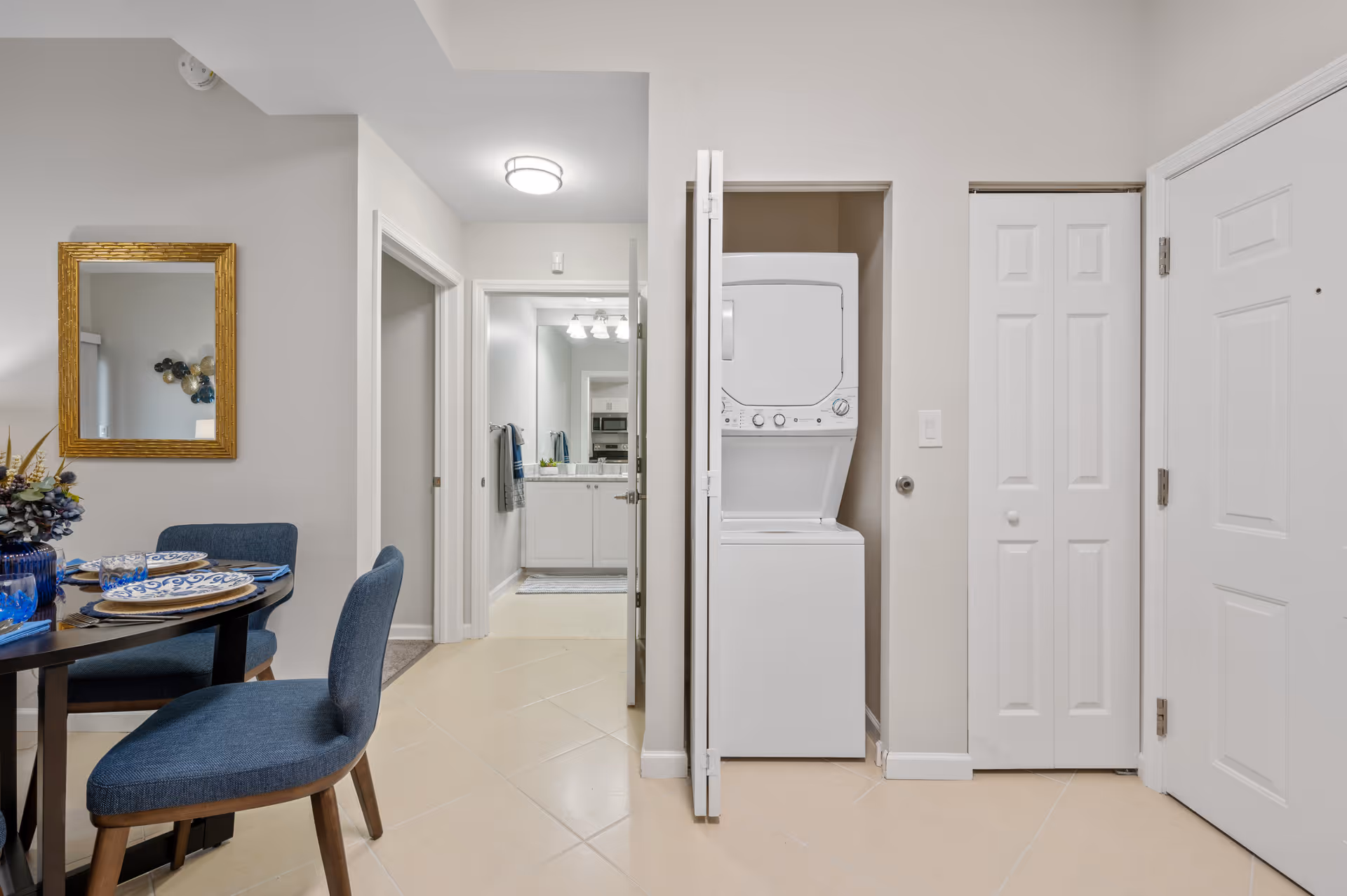 Interior view of a senior living facility showing a dining area with a round table set with plates and blue chairs. To the right, there is a closet with a stacked washer and dryer. In the background, a hallway leads to a bathroom with a sink and mirror visible.