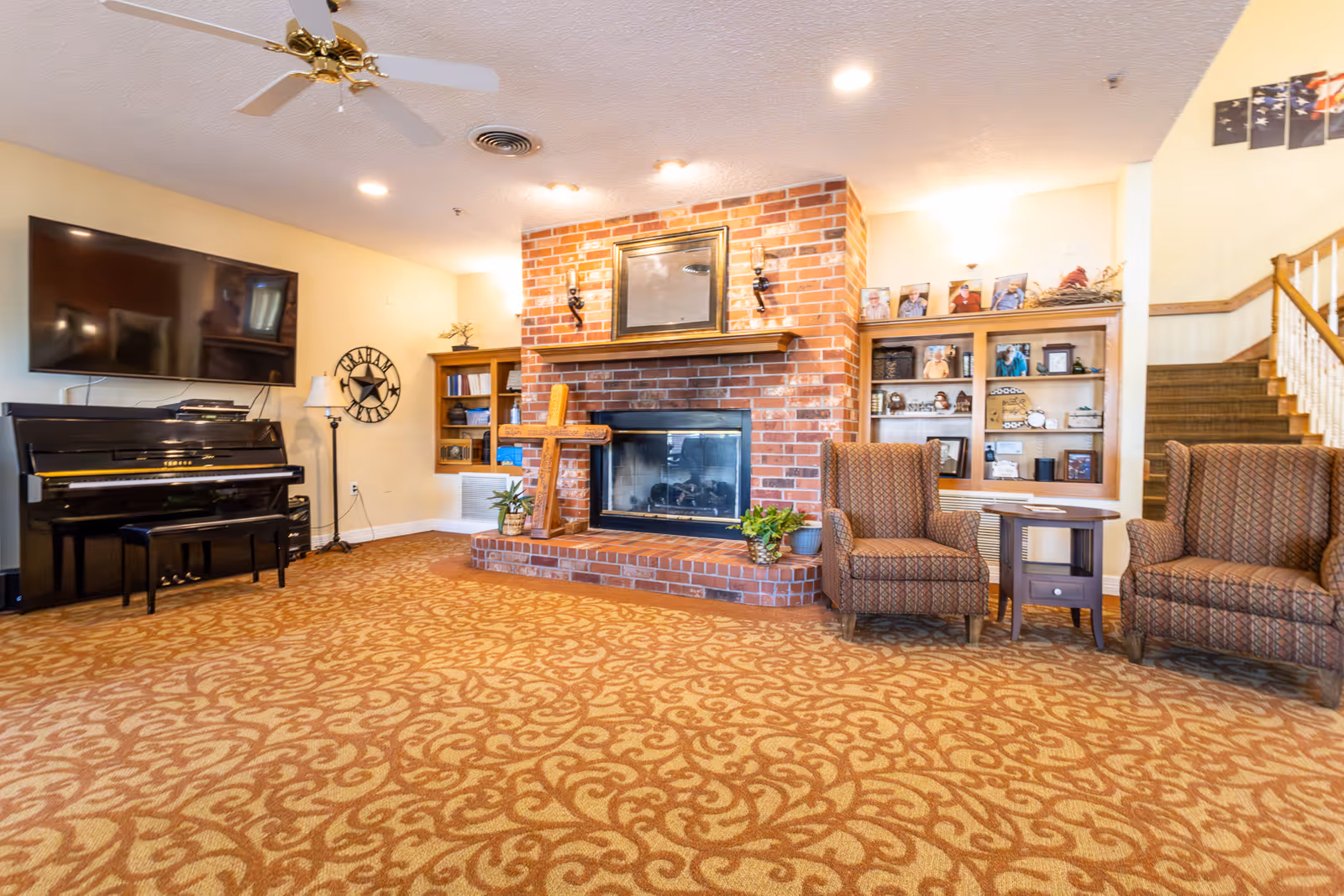 A cozy living room with a patterned carpet, a brick fireplace in the center with plants on the hearth, two patterned armchairs with a small table between them on the right, a black upright piano with a bench on the left, a large flat-screen TV mounted on the wall above the piano, built-in wooden shelves with framed photos and decorations on either side of the fireplace, and a staircase leading upstairs on the far right.