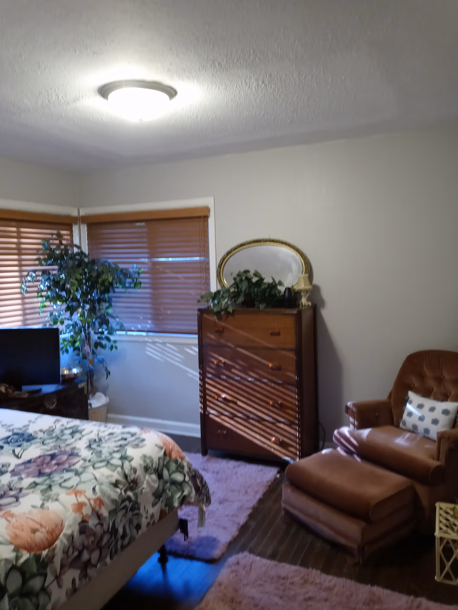 A cozy bedroom with a floral patterned bedspread, a wooden dresser with a round mirror and plants on top, a brown leather armchair with a matching ottoman, and two windows with wooden blinds. The room has a ceiling light and a soft pink rug on the wooden floor.