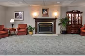 A senior living facility common area featuring a carpeted floor, a fireplace with a wooden mantle, two framed pictures above the fireplace, two wall sconces on either side of the fireplace, a wooden china cabinet on the right, two red upholstered armchairs with a small wooden table and lamp between them on the left, and several potted plants.