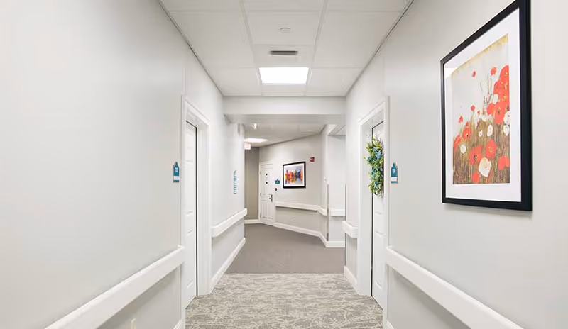 A clean, well-lit hallway in a senior living facility with light gray walls and carpeted floors. There are white doors on both sides of the hallway, one decorated with a green wreath. Handrails run along the walls, and framed artwork featuring red and white flowers is hung on the right wall. The hallway curves slightly at the end, where another framed picture is visible.