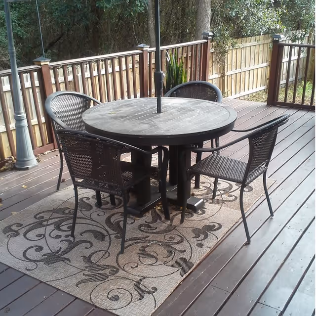 Outdoor wooden deck with a round black table and four matching black chairs arranged around it. The table has a central umbrella pole. The deck is bordered by a wooden fence and surrounded by trees and greenery. A decorative rug with a floral pattern is placed under the table and chairs.