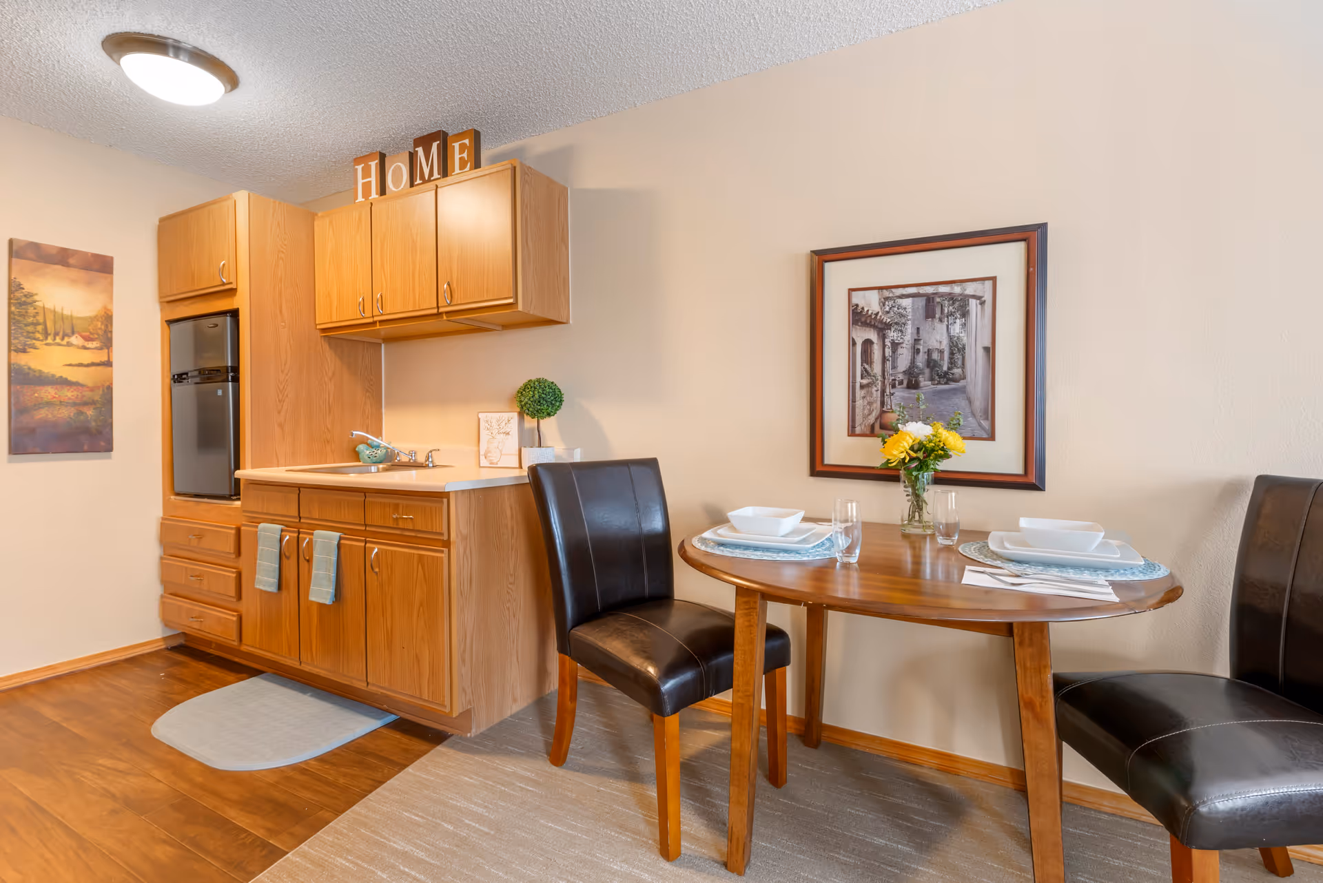 A small dining area and kitchenette in a senior living facility. The kitchenette features wooden cabinets, a small sink, and a compact refrigerator. Above the cabinets, decorative blocks spell out the word 'HOME'. The dining area has a wooden table set for two with white dishes, glasses, and a small vase of yellow flowers. Two black leather chairs are positioned at the table. A framed picture of a rustic street scene hangs on the wall above the table, and a landscape painting is on the adjacent wall.