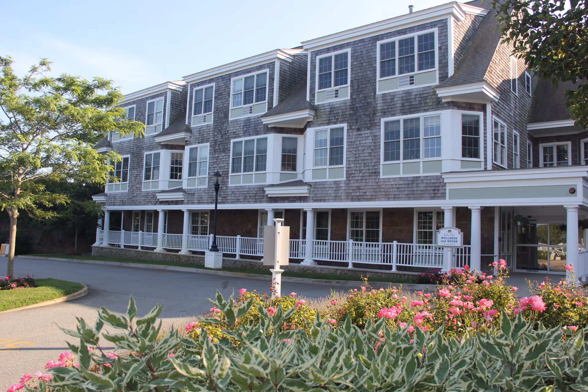 Exterior view of a multi-story senior living facility building with multiple windows, a covered entrance, and a white railing along the front porch. There are flowering bushes and greenery in the foreground and a tree to the left side of the image under a clear sky.