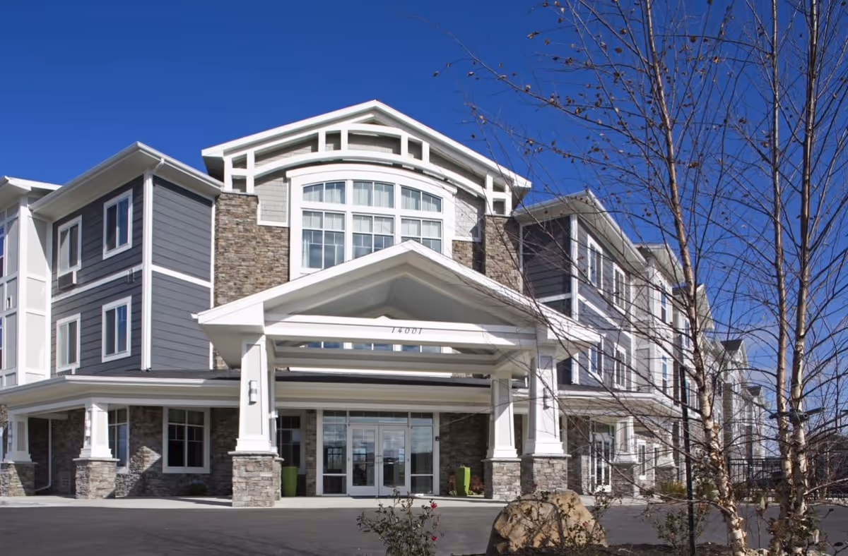 Exterior front view of a modern three-story senior living facility building with stone and gray siding, large windows, and a covered entrance under a clear blue sky.