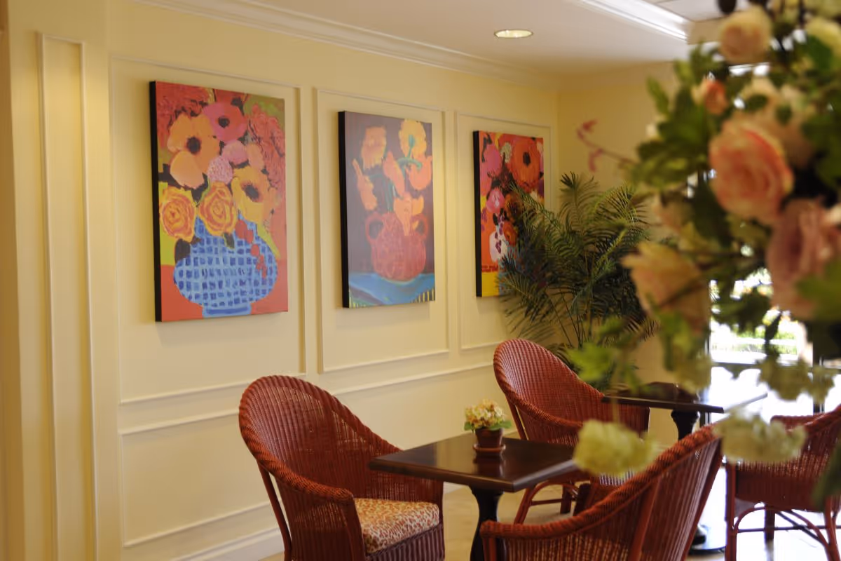 A cozy seating area with red wicker chairs around a dark wooden table, decorated with a small potted plant. The wall behind features three colorful floral paintings, and there is a large green plant in the corner. Soft lighting creates a warm and inviting atmosphere.