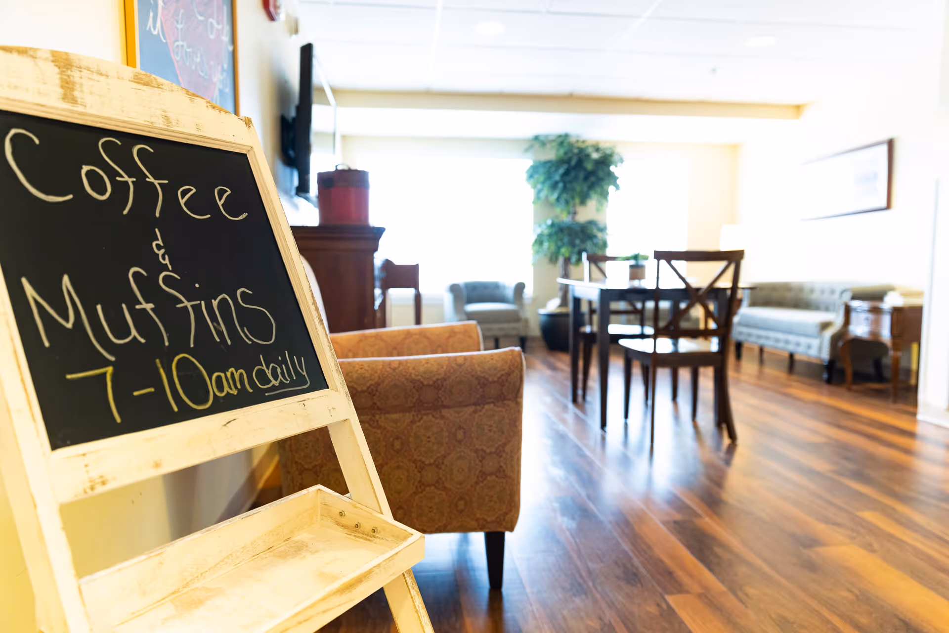 A bright and cozy senior living common area with wooden flooring, a small table with chairs, armchairs, a sofa, and a large potted plant near a window. In the foreground, there is a chalkboard sign that reads 'Coffee & Muffins 7-10 am daily.'