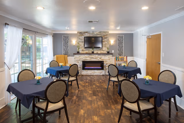 A cozy common area with four tables covered in dark blue tablecloths, each surrounded by four cushioned chairs. Small flower vases are placed on each table. The room features a stone fireplace with an electric fire and a flat-screen TV mounted above it. There are decorative wall panels and a small bookshelf next to the fireplace. Large windows with white curtains allow natural light to enter the room.