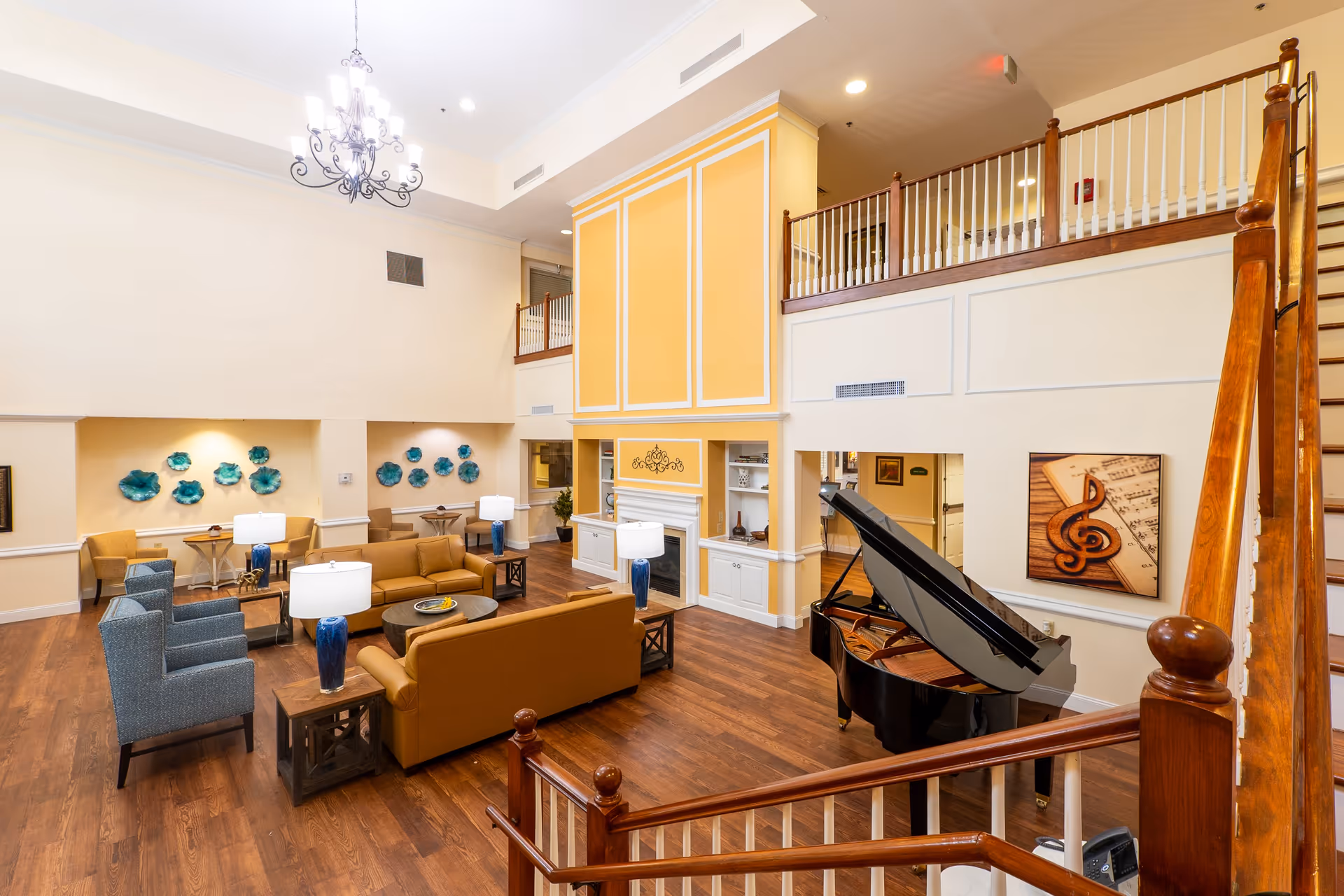 A spacious senior living facility common area with high ceilings, wooden flooring, and a large yellow and white fireplace. The room is furnished with brown sofas, blue armchairs, side tables with blue lamps, and a black grand piano. Decorative blue wall art and a musical note painting adorn the walls. A wooden staircase with white railings leads to an upper level balcony.
