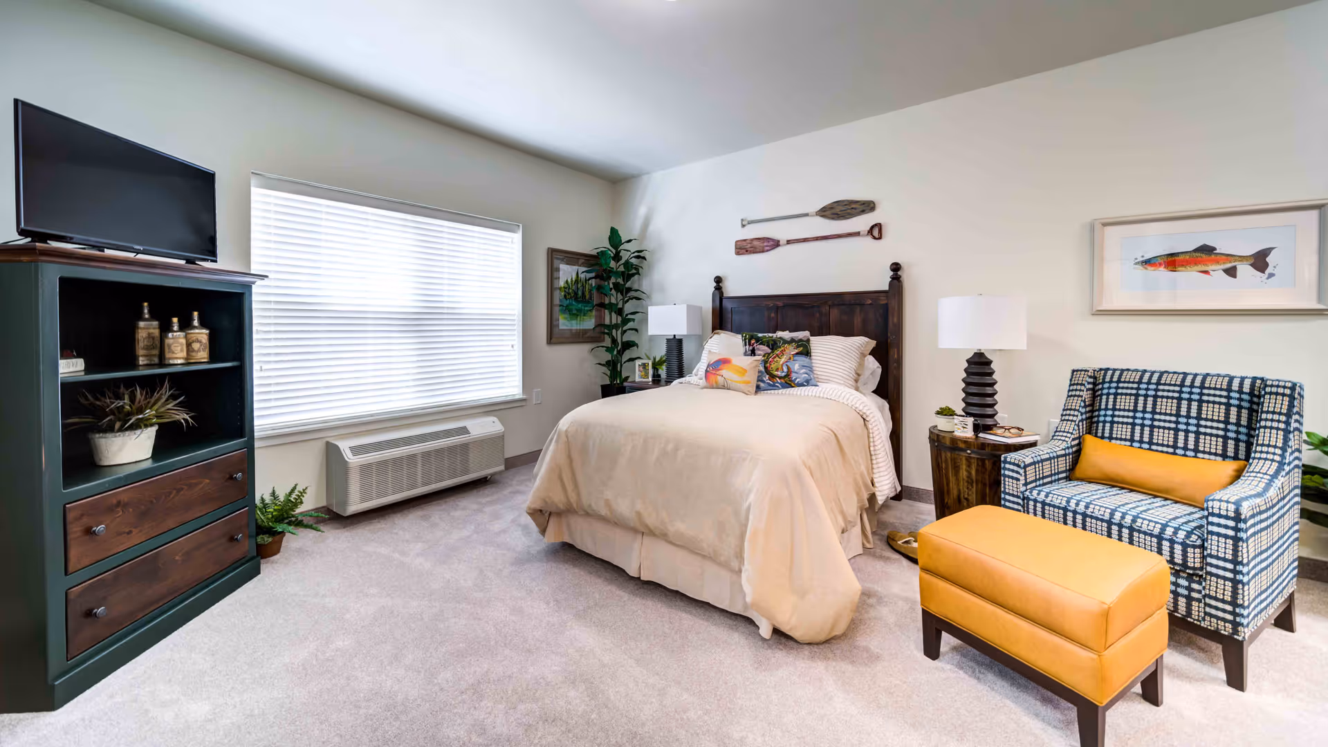 A cozy bedroom with a bed featuring beige bedding and decorative pillows, a wooden headboard with two oars mounted above it, a plaid armchair with a yellow cushion, a matching yellow ottoman, two bedside tables with lamps, a large window with blinds, a green cabinet with a TV on top, and some plants and framed artwork on the walls.