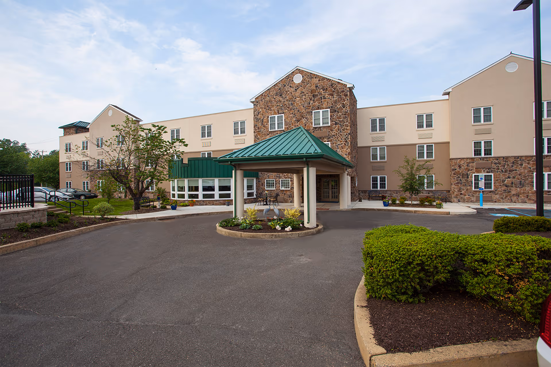 Exterior view of Commonwealth Senior Living At Willow Grove, showing a three-story building with a stone and beige facade, a green metal roof over the entrance, a circular driveway, landscaped bushes, and a few parked cars.