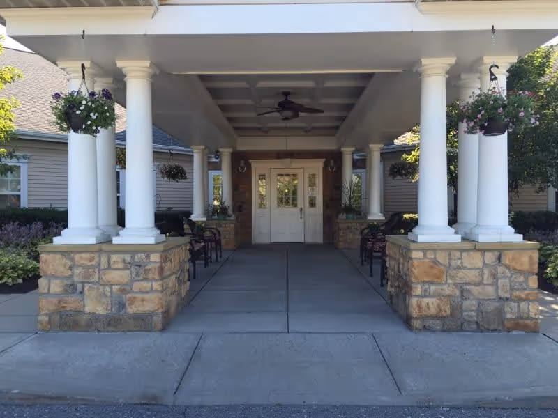 Covered entrance to a building with white double doors, supported by white columns on stone bases. Hanging flower baskets and benches line the walkway under the covered area.