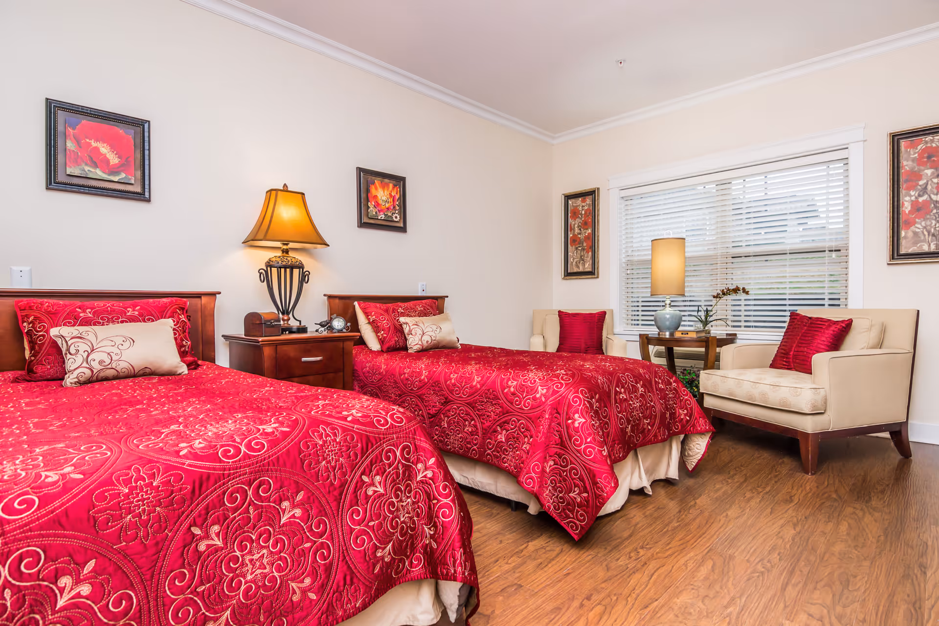 Twin bedroom with two red quilted beds, a central nightstand and lamp, two armchairs by a window, and wall art.