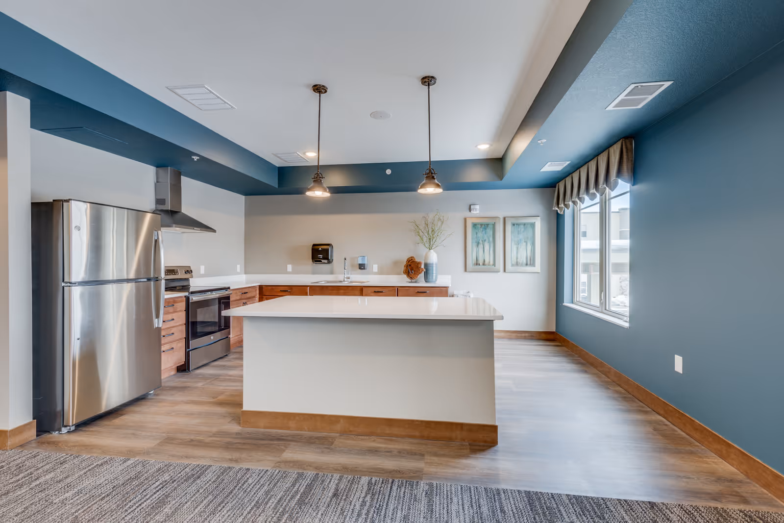 Modern communal kitchen with a large island, stainless steel refrigerator and stove, pendant lights, and a windowed seating area.