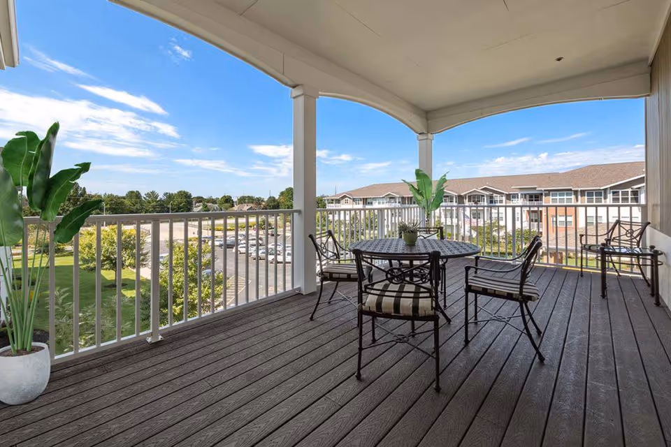 Covered outdoor balcony with a round metal table and four chairs with striped cushions. Two potted plants are placed on the balcony, and there is a view of a parking lot, trees, and a multi-story building under a blue sky with some clouds.