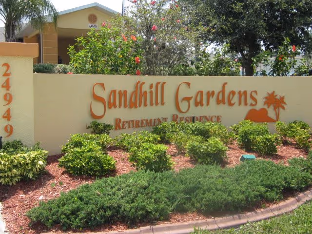 Entrance sign reading 'Sandhill Gardens Retirement Residence' mounted on a low wall surrounded by landscaped shrubs and trees.