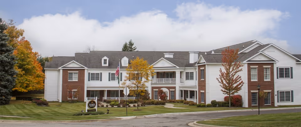 Front exterior of a two-story brick-and-white senior living building with a landscaped lawn, trees, and an American flag.