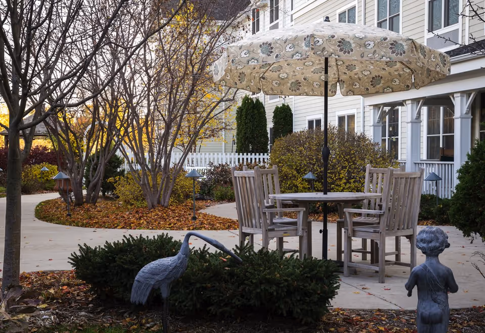Outdoor patio area at Sunrise Of Golden Valley featuring a round wooden table with four wooden chairs and a large floral-patterned umbrella. The patio is surrounded by bushes, trees with autumn leaves, a white picket fence, and a concrete pathway. There are also garden statues of a bird and a child near the patio.