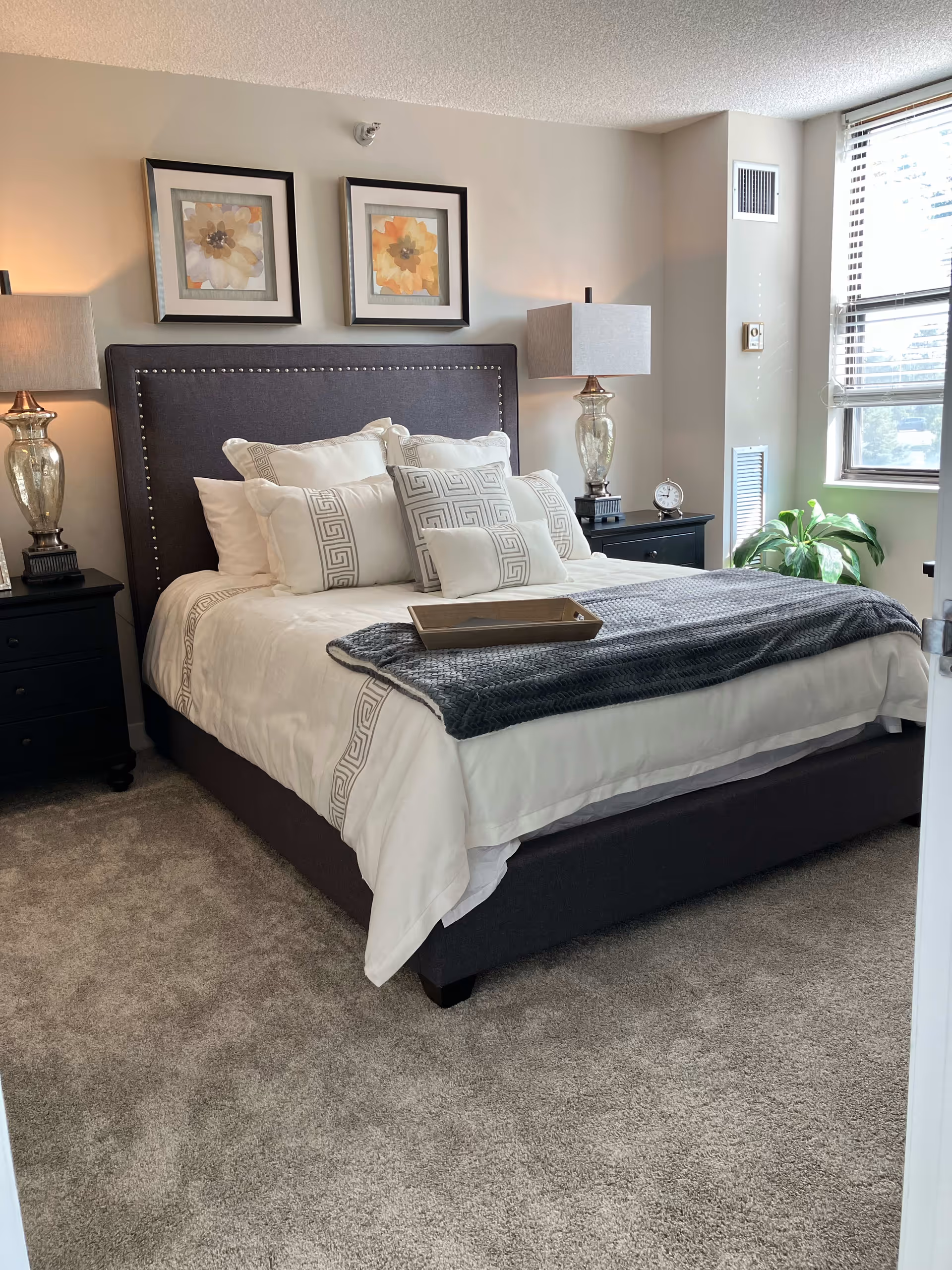 A neatly made bed with white and patterned pillows and a gray throw blanket in a bedroom. The bed has a dark upholstered headboard with nailhead trim. There are two matching bedside tables with glass lamps on each side. Two framed floral artworks hang above the bed. A window with blinds is on the right side, letting in natural light, and a green plant is placed near the window.