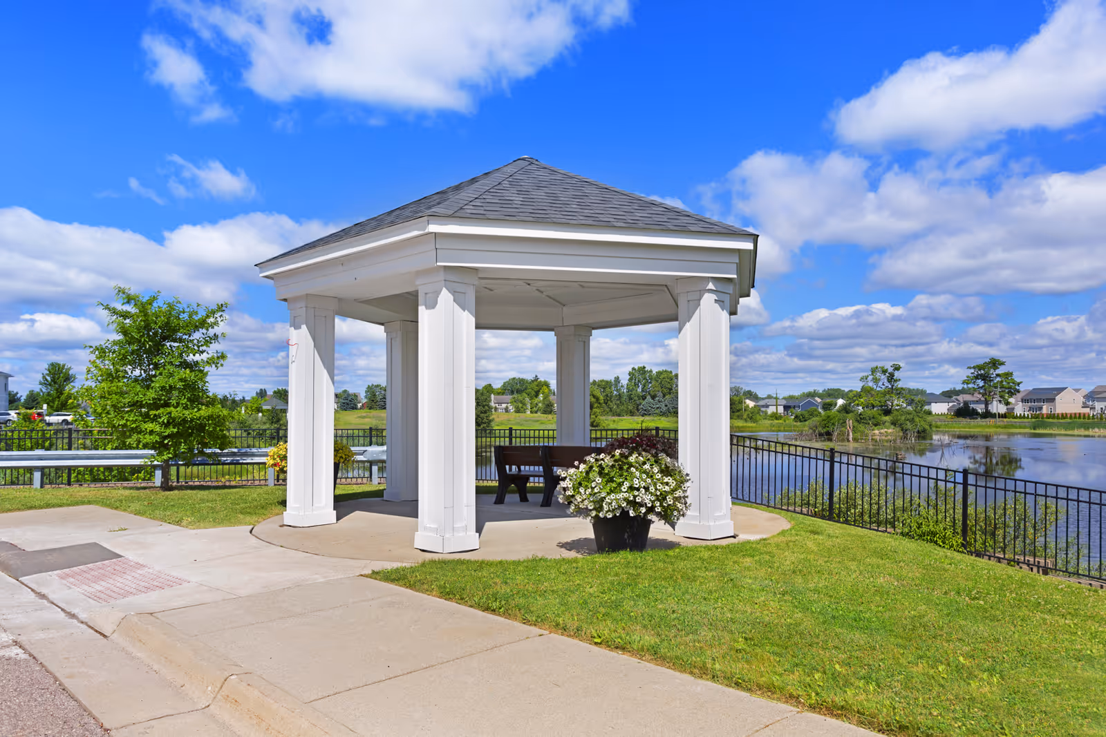 A white gazebo with a gray roof situated on a grassy area next to a paved sidewalk and a body of water. There are benches inside the gazebo and a large pot of white flowers in front. The background shows a blue sky with scattered clouds and some houses and trees across the water.