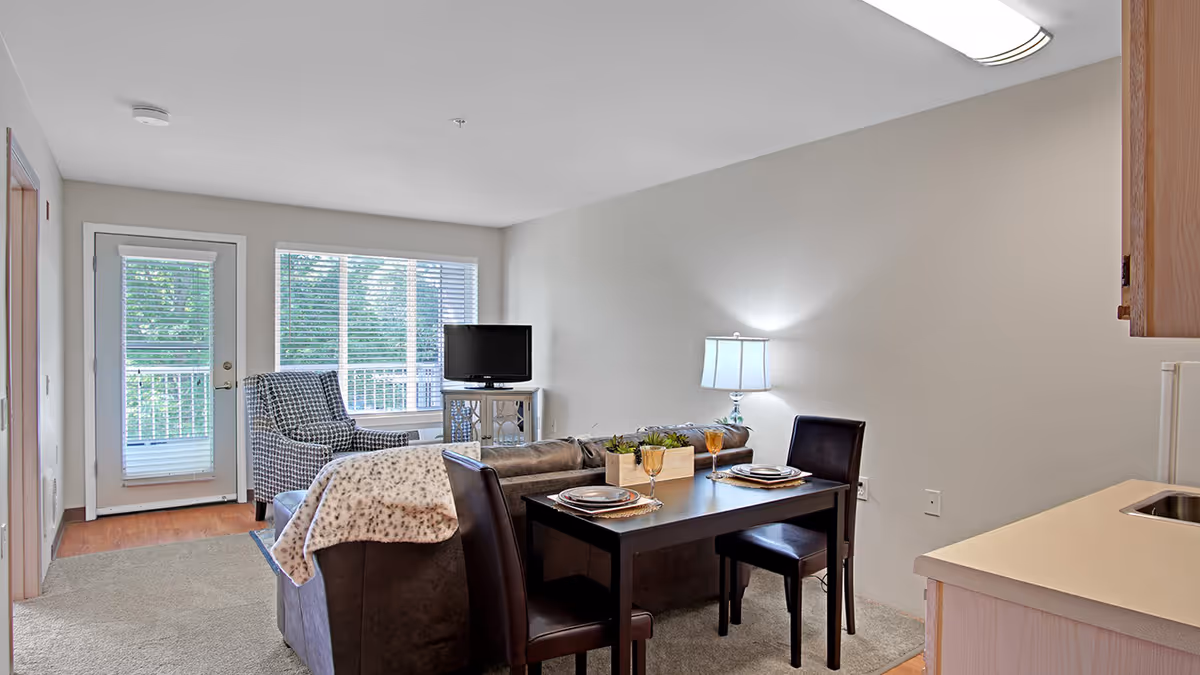 A cozy living area in a senior living facility featuring a small dining table set for two with plates and glasses, a brown leather sofa with a patterned throw blanket, a patterned armchair, a TV on a stand, a lamp on a side table, and a door and window with blinds leading to an outdoor balcony.