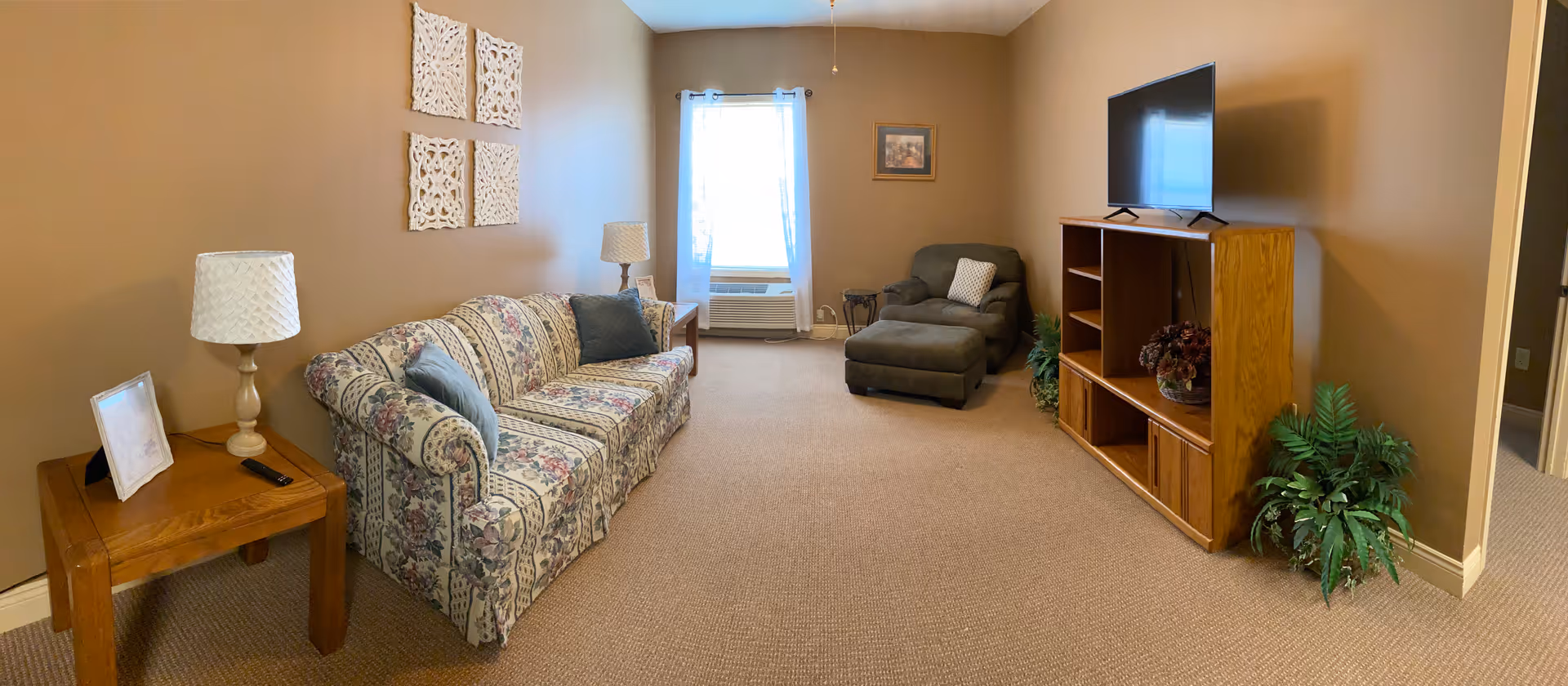 A cozy living room with beige walls and carpet. There is a floral patterned sofa with two blue pillows on the left side, accompanied by a wooden side table with a lamp and a framed photo. On the right side, there is a wooden entertainment center with a flat-screen TV on top and a decorative plant beside it. A dark gray armchair with an ottoman and a small side table are positioned near a window with sheer white curtains. Wall decorations include four white carved panels above the sofa and a framed picture above the armchair.