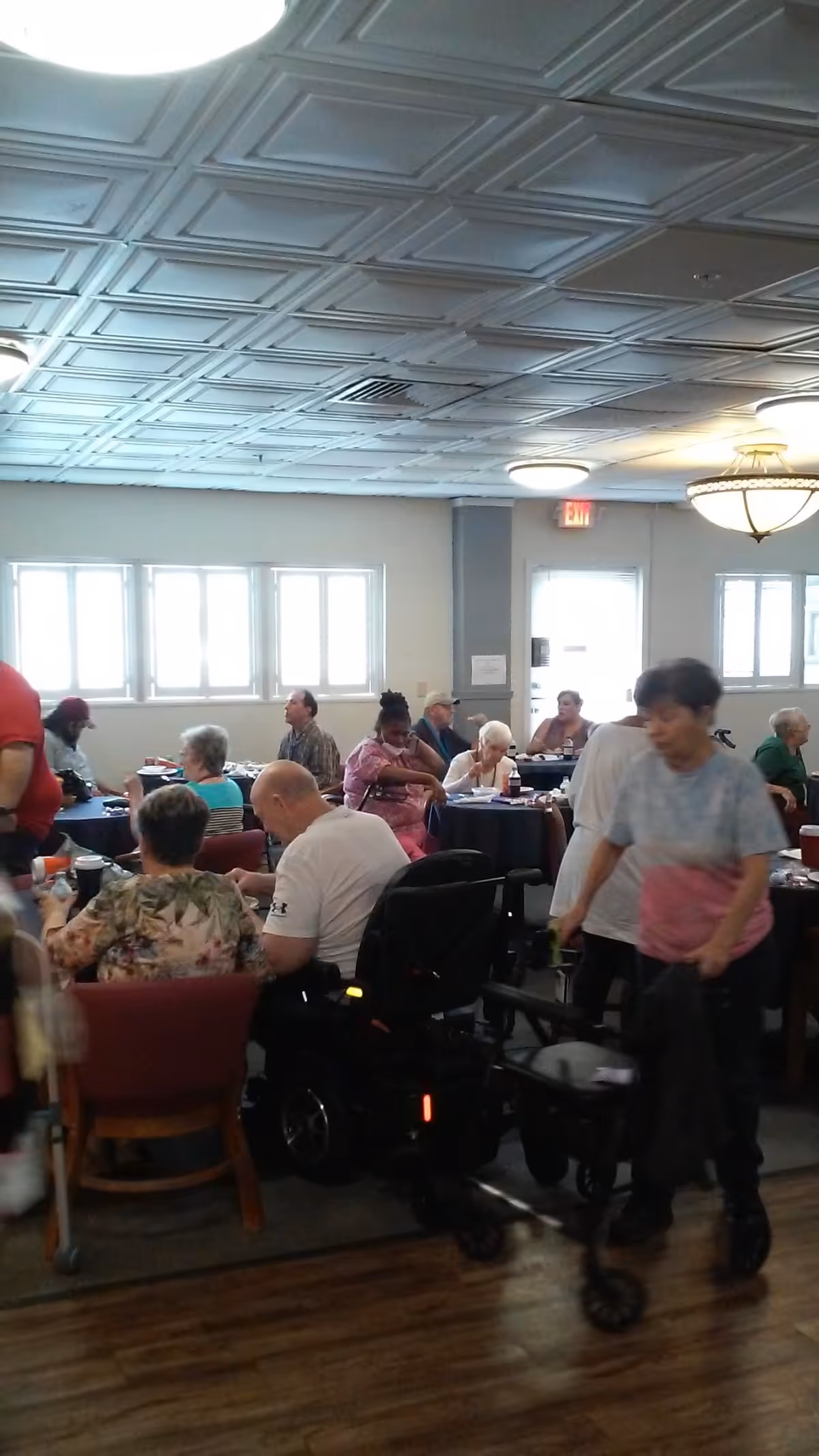 A group of elderly people sitting and socializing at tables in a well-lit room with large windows and a decorative ceiling. Some individuals are using mobility aids such as wheelchairs and walkers. The room has wooden flooring and overhead lighting fixtures.