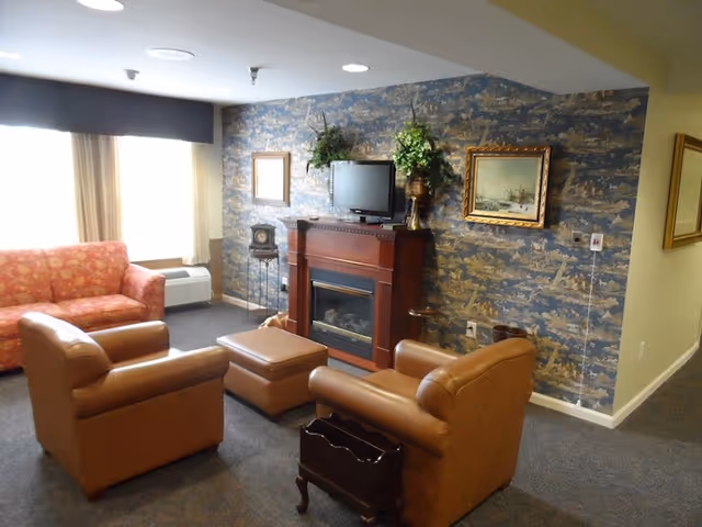 A cozy living room with two brown leather armchairs and a matching ottoman facing a wooden electric fireplace with a small TV on top. There is a red patterned sofa near a window with beige curtains. The wall behind the fireplace has blue and beige patterned wallpaper with two framed paintings and a decorative plant on the mantel.