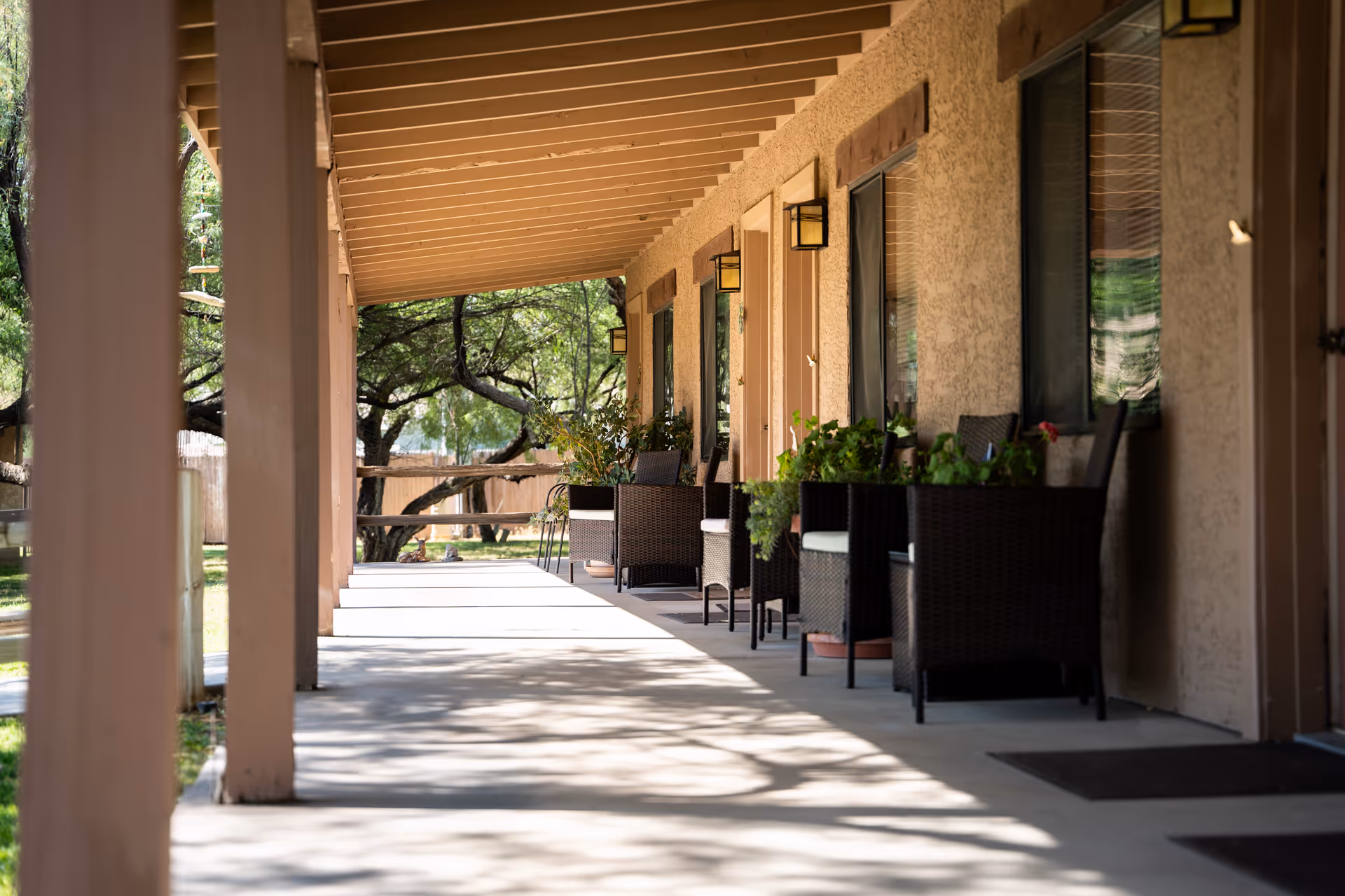 A shaded outdoor walkway at Rustic Ranch Senior Living with wicker chairs and potted plants lined up against the building's wall, under a wooden roof. Trees and greenery are visible in the background.
