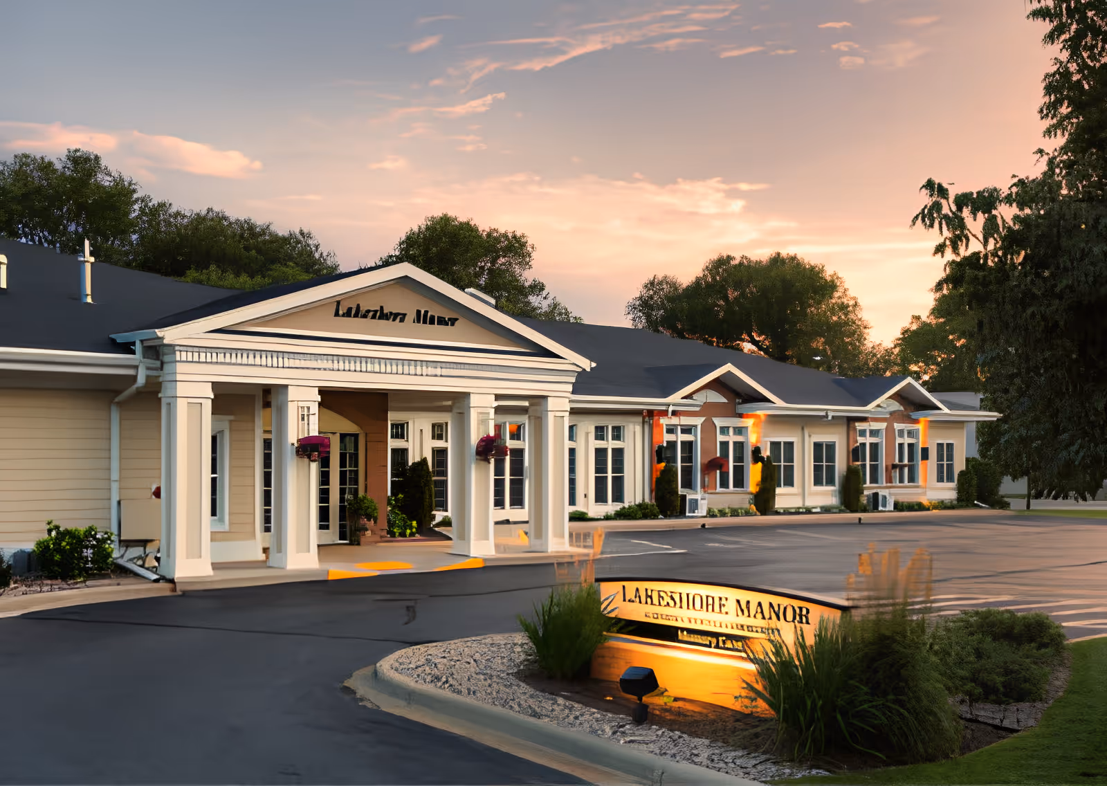 Exterior view of Lakeshore Manor building at sunset with a driveway and illuminated sign in front surrounded by greenery.