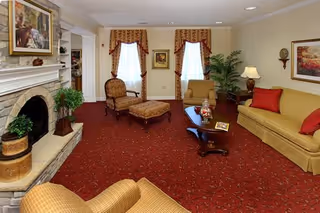 A cozy living room with a red carpet, beige walls, and traditional furniture including a beige sofa with red cushions, a matching armchair, a patterned armchair with an ottoman, and a wooden coffee table. There is a fireplace on the left with a plant on the hearth and a framed painting above it. Two windows with patterned curtains are at the back, along with a potted plant and a side table with a lamp. Additional framed artwork hangs on the walls.