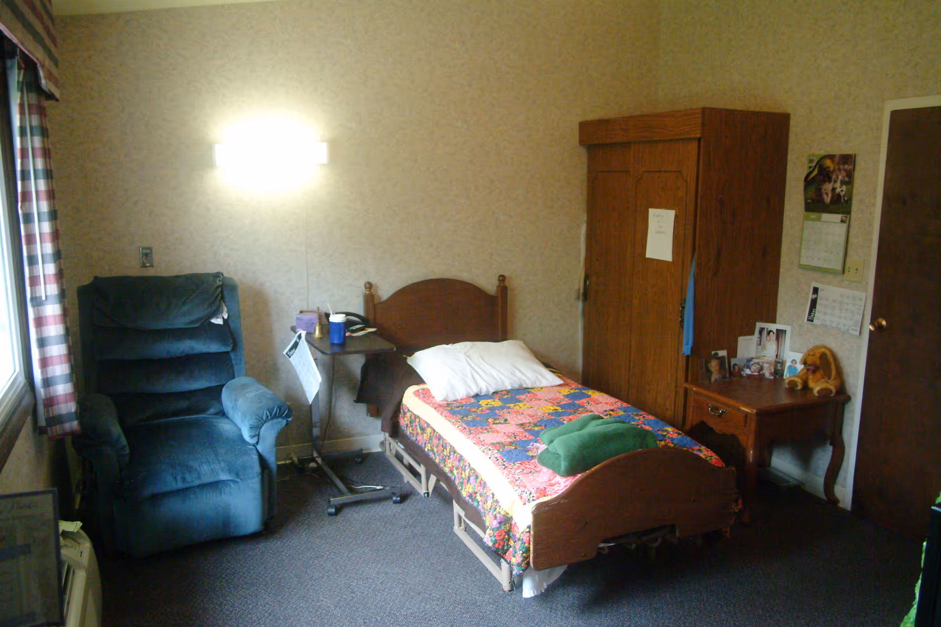 A small bedroom in a senior living facility with a single bed covered by a colorful quilt, a green blanket folded on top, and a white pillow. Next to the bed is a small wooden nightstand with framed photos and a stuffed animal. There is a wooden wardrobe against the wall and a blue recliner chair near a window with plaid curtains. A small overbed table holds a cup and some items. The walls are beige with a wall-mounted light fixture above the bed.