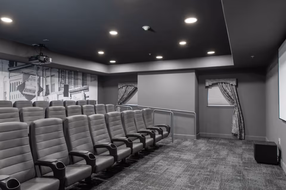 A small private theater room with tiered rows of upholstered movie seats, a ceiling-mounted projector, and a screen.