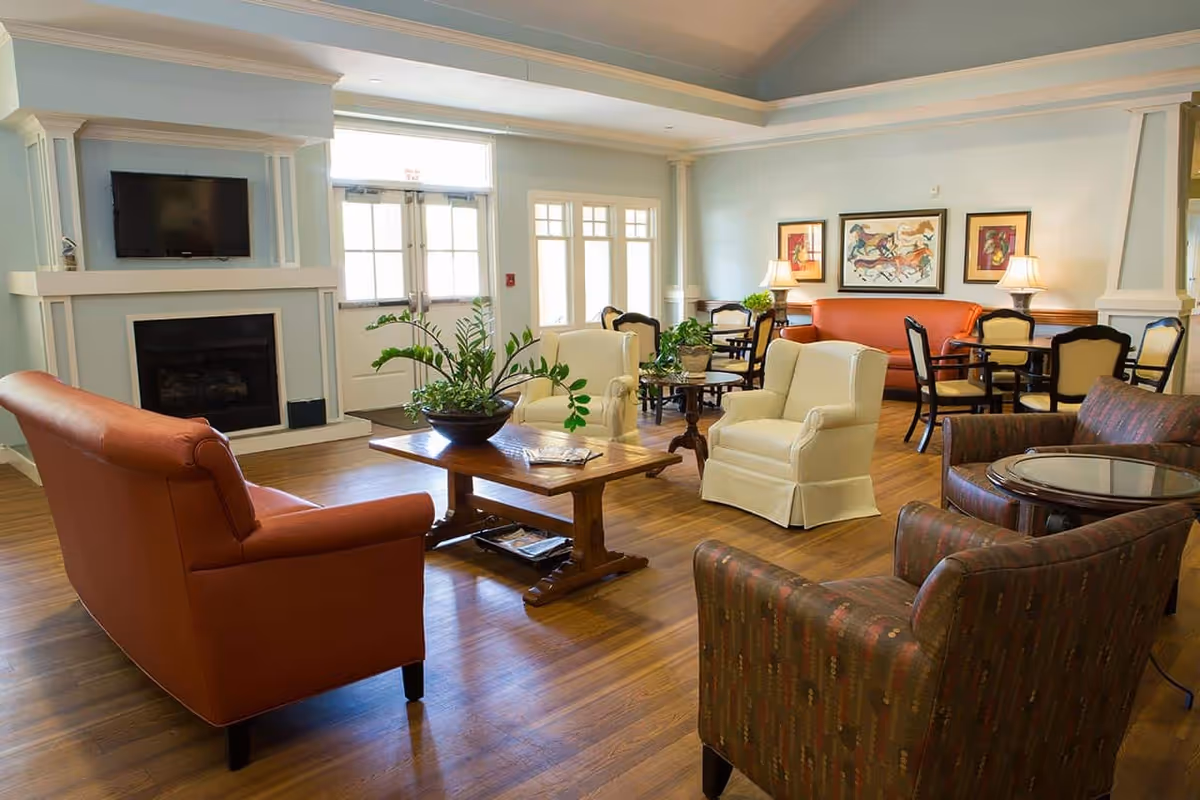 A bright and spacious living room area with hardwood floors, featuring a variety of seating options including an orange sofa, two white armchairs, and two patterned armchairs. A wooden coffee table with a plant centerpiece is in the middle. The room has a fireplace with a mounted flat-screen TV above it, large windows and glass doors letting in natural light, and framed artwork on the walls. Additional seating with tables and chairs is visible in the background.