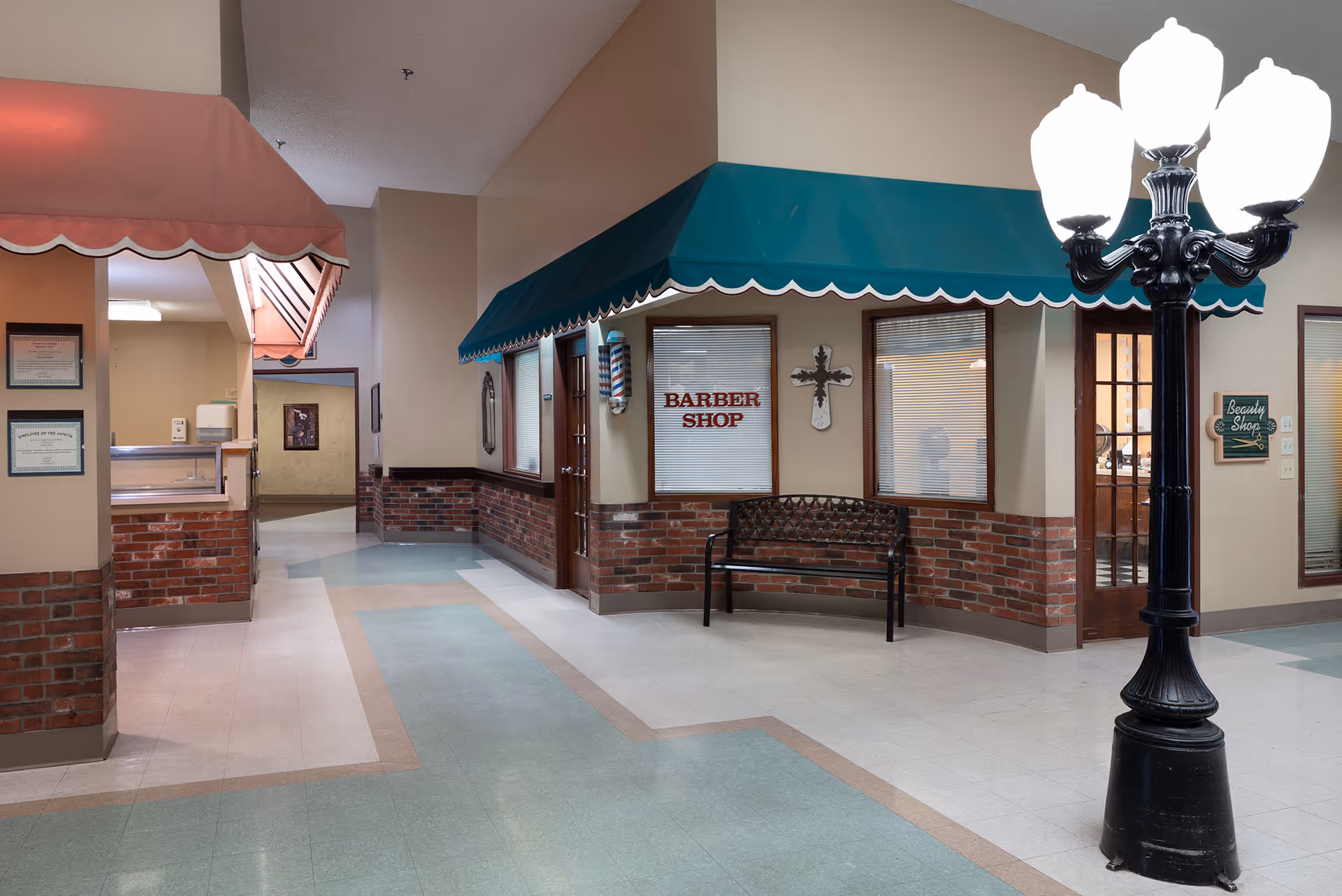 Interior hallway of a senior living facility with a barber shop and beauty shop storefronts. The barber shop has a green awning and a bench outside, while the beauty shop has a small sign next to the door. The hallway features a decorative streetlamp-style light fixture and brick half-walls with beige upper walls.