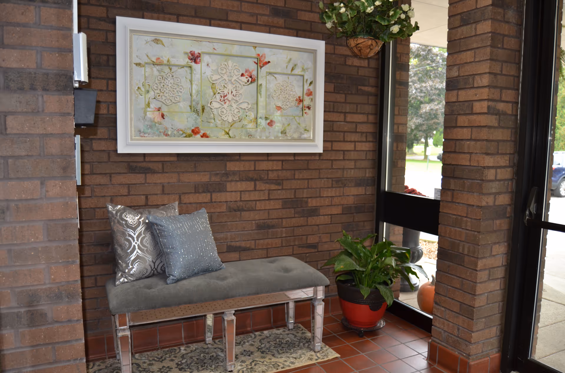 A small seating area with a gray cushioned bench adorned with two decorative pillows, placed against a brick wall. Above the bench hangs a framed artwork featuring floral and ornamental designs. To the right, there is a glass door and window showing an outdoor view with greenery. A potted plant sits on the floor near the door.