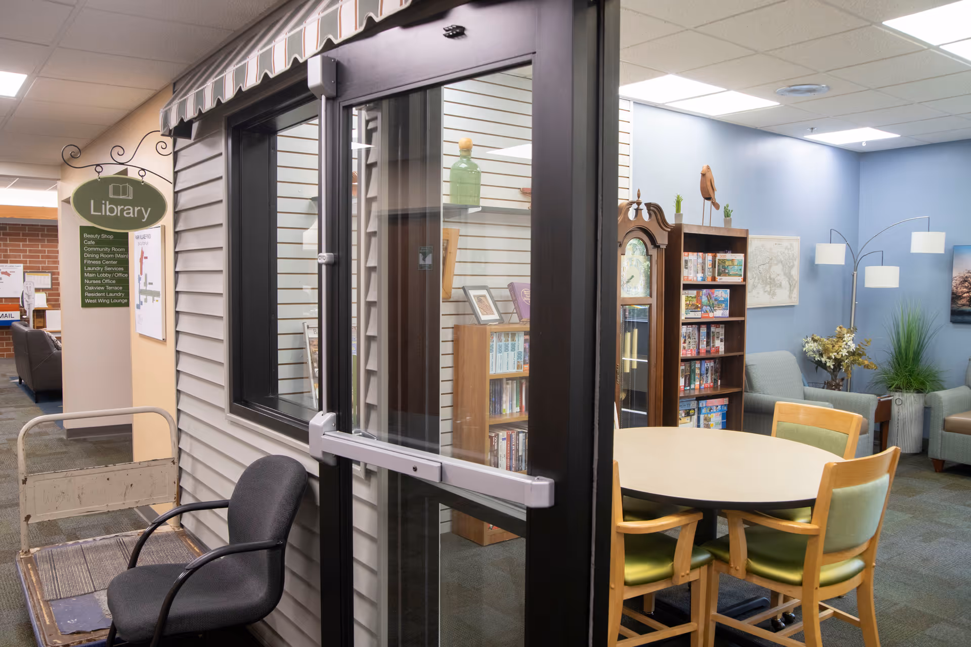 Interior view of a library area in a senior living facility with a round table surrounded by four chairs, a tall grandfather clock, bookshelves filled with books and games, a blue wall with framed pictures, and a modern floor lamp. A glass door with a black frame is in the foreground, and a sign indicating the library and other facility areas is visible on the left.