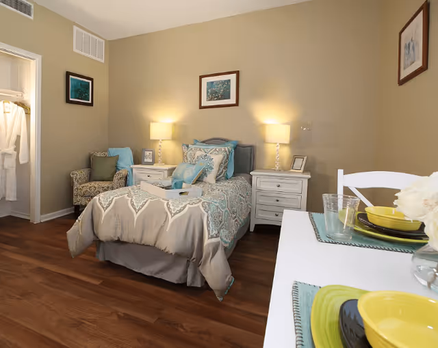 A cozy bedroom in West Fork Village featuring a single bed with patterned bedding and multiple pillows, two white nightstands with lamps on each side of the bed, a patterned armchair in the corner, framed artwork on the walls, and a small dining table set with yellow and black dishes and clear glasses. The room has wooden flooring and beige walls.
