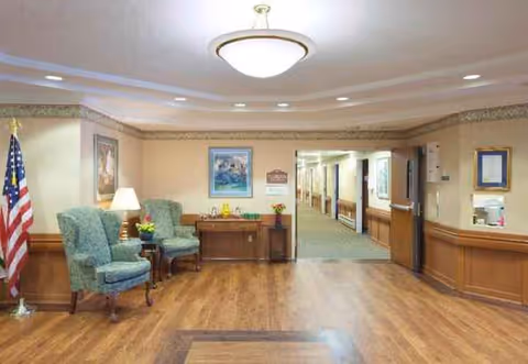 Well-lit senior living lobby with green upholstered chairs, an American flag, and a hallway.